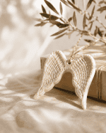 Cream ceramic angel wings ornament displayed on soft linen with olive branches in warm natural light, styled as a meaningful keepsake gift.