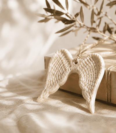 Cream ceramic angel wings ornament displayed on soft linen with olive branches in warm natural light, styled as a meaningful keepsake gift.