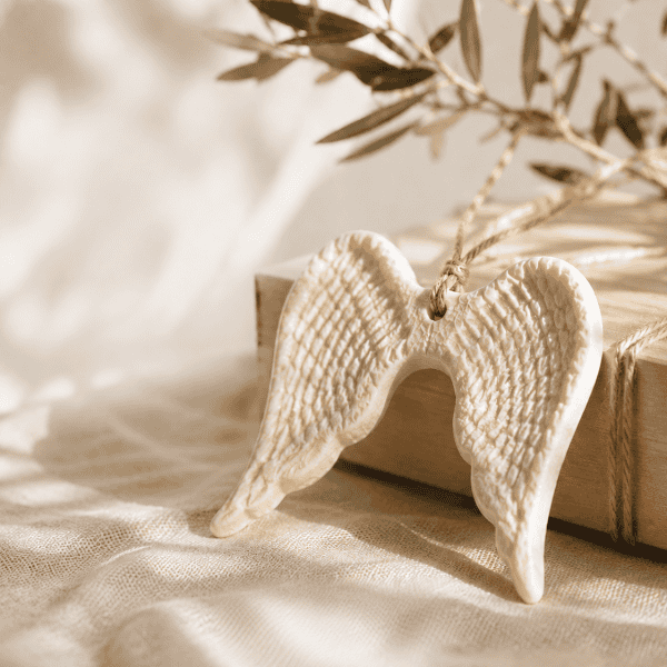 Cream ceramic angel wings ornament displayed on soft linen with olive branches in warm natural light, styled as a meaningful keepsake gift.