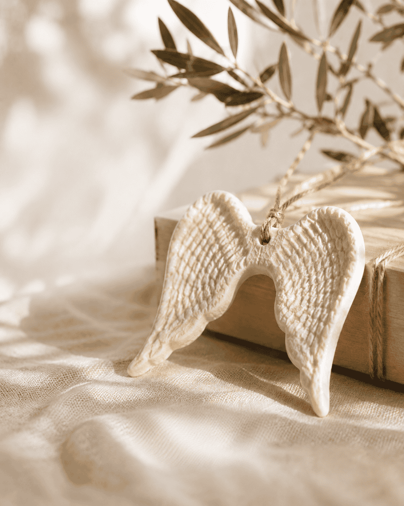 Cream ceramic angel wings ornament displayed on soft linen with olive branches in warm natural light, styled as a meaningful keepsake gift.