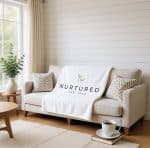Soft beige sofa with decorative pillows, a white blanket with "Nurtured" logo, a coffee cup, and a p.