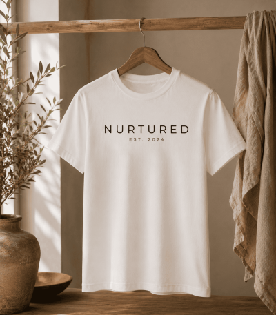 Minimalist white Nurtured graphic t-shirt hanging on a rustic wooden rod in a warm neutral interior with natural textures and soft inspired styling.