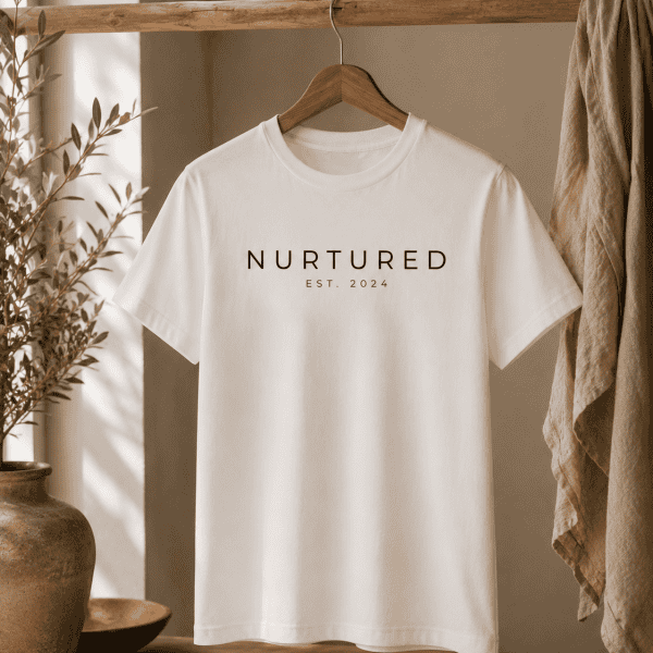 Minimalist white Nurtured graphic t-shirt hanging on a rustic wooden rod in a warm neutral interior with natural textures and soft inspired styling.