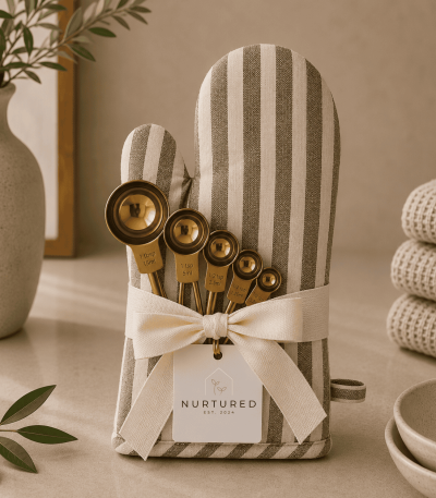 Luxury kitchen gift set featuring a striped cotton oven mitt wrapped with a cream ribbon and polished gold stainless steel measuring spoons in a warm neutral kitchen setting.